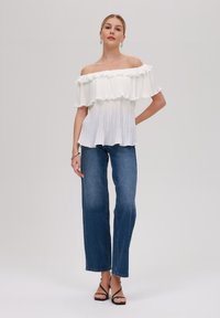 Weiße schulterfreie Bluse mit rüschierten Schichten und Falten, kombiniert mit hochgeschnittenen blauen Jeans und schwarzen Riemchensandalen.