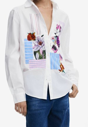Chemise blanche boutonnée avec des patchs floraux colorés et un motif Minnie Mouse, portée avec un jean bleu, montrée du cou aux hanches.