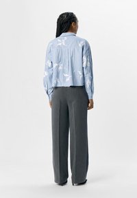 Camicia a righe blu chiaro con motivo floreale bianco, linea rilassata e maniche lunghe. Abbinata a pantaloni grigi a gamba larga, che mostrano un look casual.