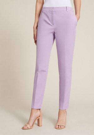 Vrouw draagt op maat gemaakte lila enkelhoge broek met voorplooi en nudekleurige sandalen met hoge hakken, staand tegen een neutrale achtergrond.