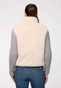 Kremkrāsas mākslīgā pūka veste ar augstu apkakli, apvienota ar pelēkiem adītiem piedurknēm un ziliem džinsu biksēm. Vestes virsma ir teksturēta un mīksta.