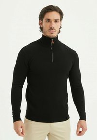 Pull en col zip noir au design ajusté, avec des manches longues et un tissu texturé. La fermeture éclaire est équipée d'un tirant en cuir marron.