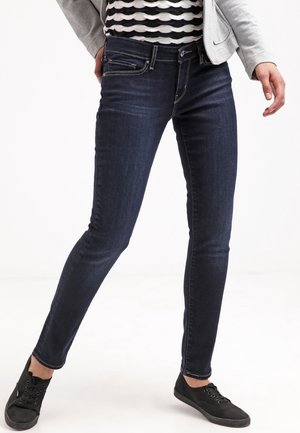 Nainen pukeutuneena tummansinisiin skinny-farkkuihin, mustiin tennareihin, raidalliseen paitaan ja vaaleanharmaaseen takkiin, seisoo jalat leveästi erillään valkoisella taustalla.