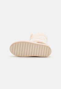 Beige pelsete støvel med en teksturert overflate, snøre detaljer og en riftet gummisåle for grep. Designet er mykt og sylindrisk i form.