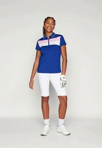 Μπλε γκολφ πόλο με ροζ και λευκή ριγέ σχέδιο στο στήθος, συνδυασμένο με λευκά σορτς και λευκά παπούτσια γκολφ. Περιλαμβάνει και γάντι γκολφ.