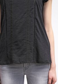 Czarna bluzka z krótkim rękawem z koronkowymi wykończeniami wzdłuż środka i przy rękawach, wykonana z miękkiego, lekkiego materiału.