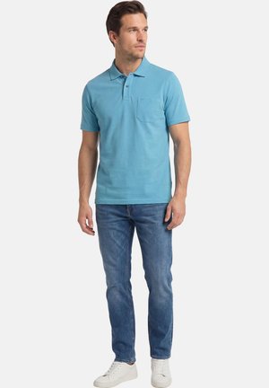 Mann trägt hellblaues Poloshirt, blaue Jeans und weiße Turnschuhe, steht und schaut seitlich vor einem einfarbigen Hintergrund.