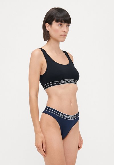 Zwarte sport BH met brede banden en een witte "Emporio Armani" logo band, gecombineerd met een blauwe string met vergelijkbaar logo en streepdetaillering.