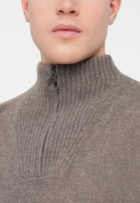 Κοντινό πλάνο ενός ατόμου που φοράει έναν γκρι ριγωτό πλεκτό πουλόβερ με ψηλό γιακά και δαχτυλίδι μ zipper με το λογότυπο "Barbour."