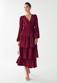 Burgundowa maksikowa sukienka z falbanami, długimi rękawami, dekoltem w serek, dopasowaną talią i miękkiego materiału. Stylizowana z białymi sandałami na ramiączkach i małą torebką.