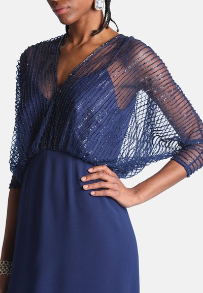 Femme portant une robe bleu marine avec une superposition transparente et pailletée sur le haut du buste et les manches, posant avec une main sur la hanche.