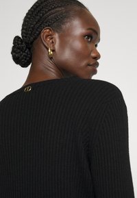 Γυναίκα με χτενίσμα κοτσίδα σε σχήμα σφιχτού σφιξίματος, φορά χρυσό σκουλαρίκι κρίκο και μαύρη ριμπ μπλούζα με μικρό χρυσό λογότυπο στην πάνω πλάτη.