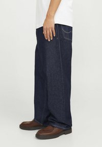 Mörkblå oversized denimjeans med en avslappnad passform, som har gula sömmar och sidofickor, i kombination med bruna avslappnade skor.