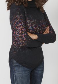 Černý rolák s barevnými skvrnami, pletenou texturou a mírně zakřiveným lemem. V kombinaci s modrými džíny.