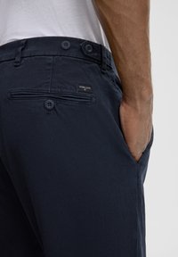 Pantaloni in cotone navy con frontale piatto, due tasche posteriori e patch con logo. Presentano bottoni in vita e tasca a filetto laterale.