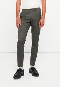 Bărbat îmbrăcat în pantaloni gri slim-fit, șosete albe, încălțăminte neagră cu șireturi și un top alb fără mâneci, pe un fundal alb uniform.