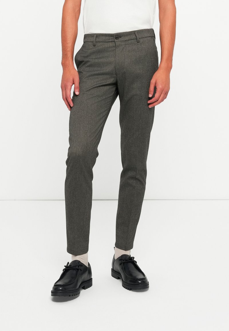 Bărbat îmbrăcat în pantaloni gri slim-fit, șosete albe, încălțăminte neagră cu șireturi și un top alb fără mâneci, pe un fundal alb uniform.