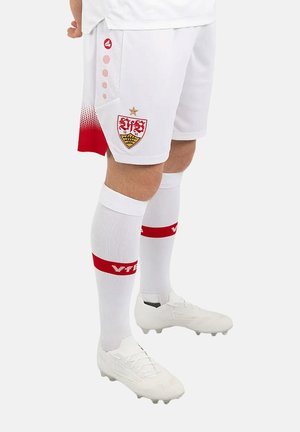 Gambe maschili che indossano pantaloncini da calcio bianchi con dettagli rossi, calzettoni bianchi con bande rosse e scarpe da calcio bianche su uno sfondo semplice.