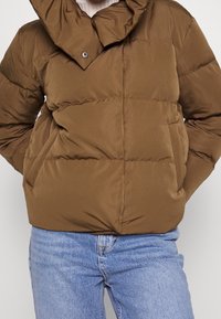 Chaqueta de plumas marrón con alto cuello, con cierre de botones, diseño acolchado y bolsillos laterales, combinada con jeans de color azul claro.