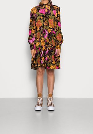 Kobieta w czarnej sukience do kolan z żółtymi i różowymi kwiatowymi wzorami oraz brązowych sneakersach za kostkę z białymi sznurowadłami.