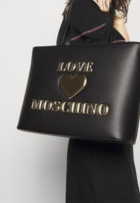 Musta nahkainen käsilaukku, jossa on kultametrinen "LOVE" ja sydämen emblem, ja "MOSCHINO" on kaiverrettu alle samankaltaisella fontilla.