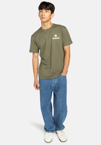 Olívovo zelené bavlnené tričko s krátkymi rukávmi a okrúhlym výstrihom, s bielym logom na ľavom hrudi, v kombinácii s voľnými modrými džínami.