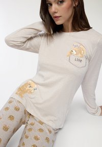 Camicia a maniche lunghe di un beige chiaro con grafica di un orso che indossa un fiocco e il testo "LOVE". Pantaloni da pigiama beige coordinati con un motivo ripetuto di orsi.