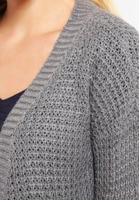 Grå stickad cardigan med en strukturerad öppen väv och ribbad halsringning. Tyget är lätt och visar upp intrikata detaljer i stickningen.