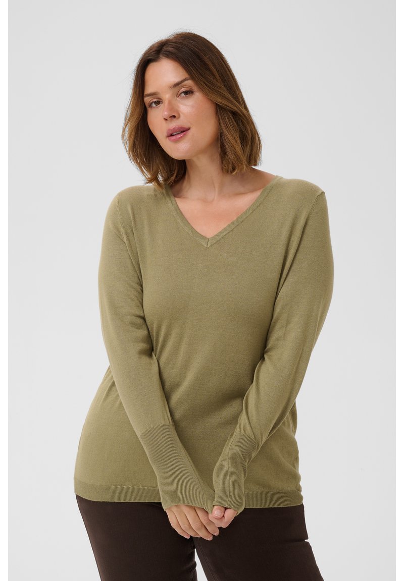 Femme aux cheveux bruns mi-longs, portant un pull col en V vert olive clair et un pantalon foncé, posant avec les mains jointes devant elle.