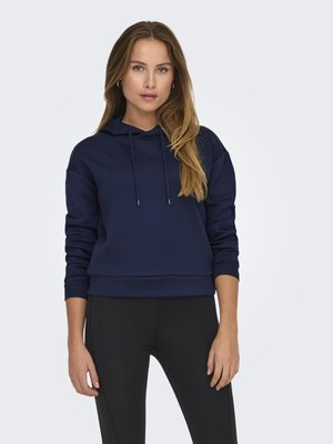 Femme aux longs cheveux portant un sweat à capuche bleu marine et un pantalon noir, debout devant un fond clair uni.