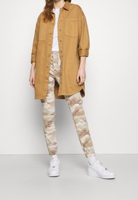 Paita pitkähihaisena camel-värissä, jossa on kaksi rintataskua, yhdistettynä istuviin vaaleanbeigeihin camo-housuihin ja valkoisiin lenkkikenkiin.