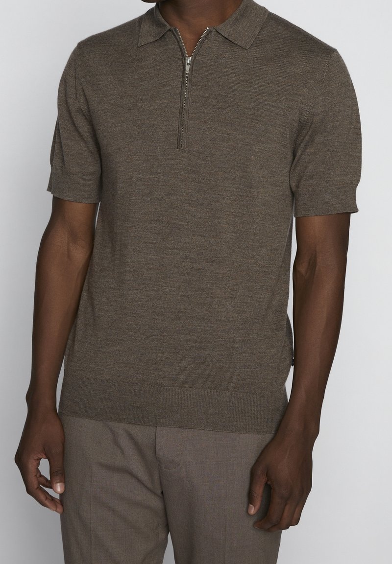 Homme portant un polo en maille marron à manches courtes avec une fermeture éclair devant, associé à un pantalon marron assorti sur un fond uni.