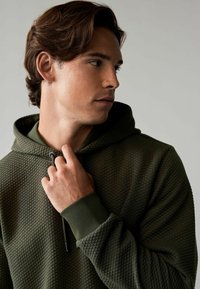 Olijfgroene hoodie van een gestructureerde stof met een diamantpatroon. Voorzien van een trekkoordsluiting en geribbelde manchetten. Close-up van het model.