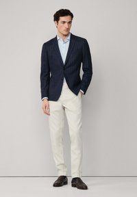 Námořnický blazer s proužky, světle modrá košile, béžové kalhoty a tmavě hnědé boty. Ušitý design se strukturovaným tvarem a kontrastními barvami.
