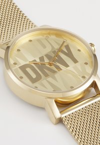 Orologio dorato con cinturino a rete, con un quadrante texturizzato che mostra in modo prominente il logo "DKNY". Indicatori delle ore e lancette minimalisti.