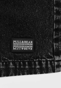 Tissu en denim noir avec une étiquette rectangulaire cousue indiquant "PULL&BEAR DENIM GOODS SUPPLIER" en lettres majuscules blanches.