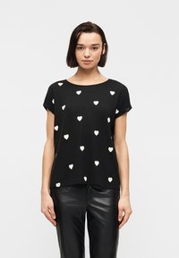 Γυναίκα που φοράει μαύρο κοντομάνικο μπλουζάκι με λευκά μοτίβα καρδιών και μαύρο δερμάτινο παντελόνι, στέκεται μπροστά σε μια απλή λευκή στάση.