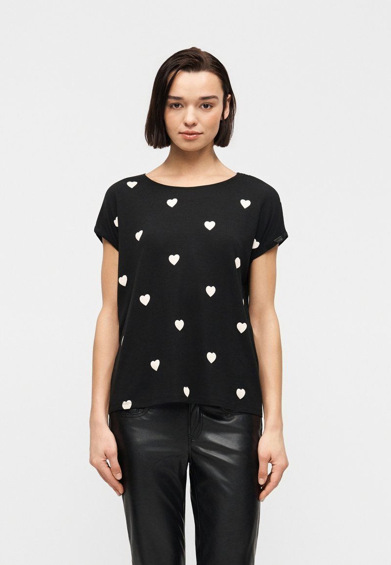 Γυναίκα που φοράει μαύρο κοντομάνικο μπλουζάκι με λευκά μοτίβα καρδιών και μαύρο δερμάτινο παντελόνι, στέκεται μπροστά σε μια απλή λευκή στάση.