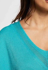 Haut turquoise en maille avec un décolleté en V orné d'une fine bordure en maille ajourée à motifs sur les bords. Texture douce avec une apparence légère.