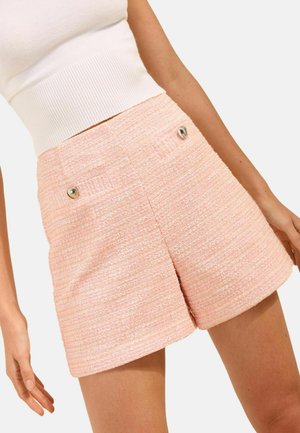 Woman wearing high-waisted, textured pink shorts with metallic buttons and a white sleeveless ribbed crop top.