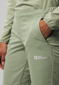 Pantaloni da esterno verde chiaro con tasca con cerniera e logo Jack Wolfskin bianco. Tessuto liscio con vestibilità adattata.