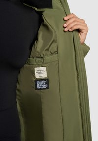 Oliivroheline polsterdatud jope, mille siseküljel on must silt; omab siledat tekstuuri, õmmeldud silte ja tugevat disaini.