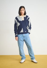 Ναυτικό και γκρι πουλόβερ με αφηρημένα σχέδια, μπεζ γιακά και τρία κουμπιά, συνδυασμένο με ανοιχτό μπλε παντελόνι με φαρδιές γραμμές και λευκά αθλητικά παπούτσια.