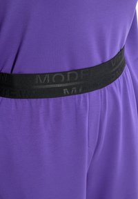 Lila Stoff mit einer sanften Textur, ausgestattet mit einem breiten schwarzen Gummibund, der mit dem Text "MODERN VIEW" geprägt ist. Einfaches, figurbetontes Design.