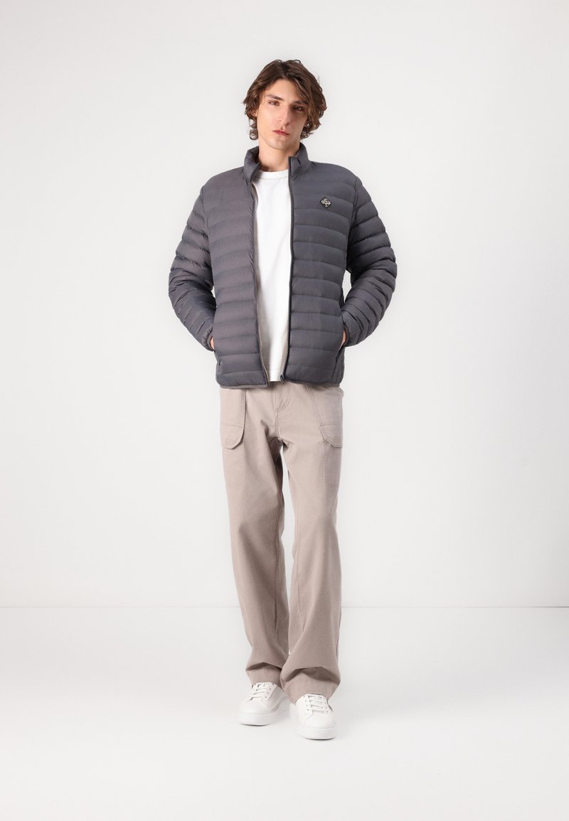 Veste grise rembourrée avec des coutures horizontales, col montant, fermeture éclair à l'avant et logo sur la poitrine, portée sur un t-shirt blanc et un pantalon beige.