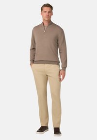 Beiger Sweater mit Reißverschlusskragen aus weichem Strickstoff, kombiniert mit beigen Hosen. Das Model trägt dunkle Sneaker und steht vor einem schlichten weißen Hintergrund.