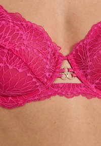 Soutien-gorge di pizzo rosa brillante con motivi floreali, dettaglio in anello metallico al centro, indossato su una pelle chiara.