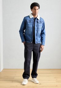 Jeansjacke mit einem weißen Lammfellkragen, zwei Brusttaschen, über einem schwarzen Hemd getragen und kombiniert mit dunklen Cargo-Hosen und weißen Sneakers.