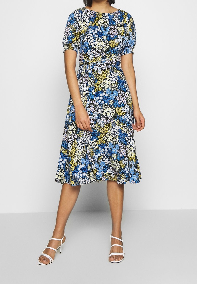 Květinové šaty s dobře padnoucím pasem, krátkými nabíranými rukávy a délkou ke kolenům. Obsahují modré, žluté a bílé vzory na černém pozadí.