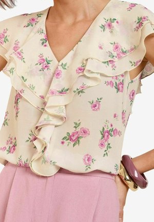 Mujer con blusa floral color crema con mangas con volantes y pantalones rosas, complementada con grandes pulseras doradas y moradas.